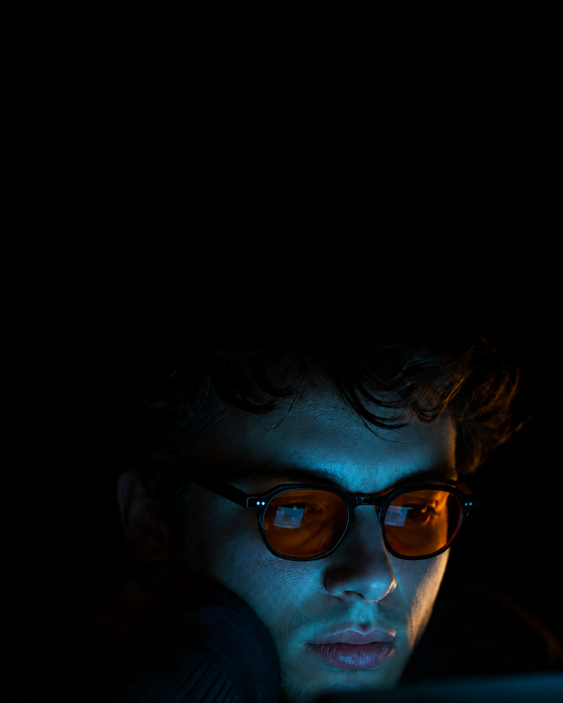 Homem no escuro usando óculos de lente âmbar Biohack, rosto iluminado pela luz azul da tela